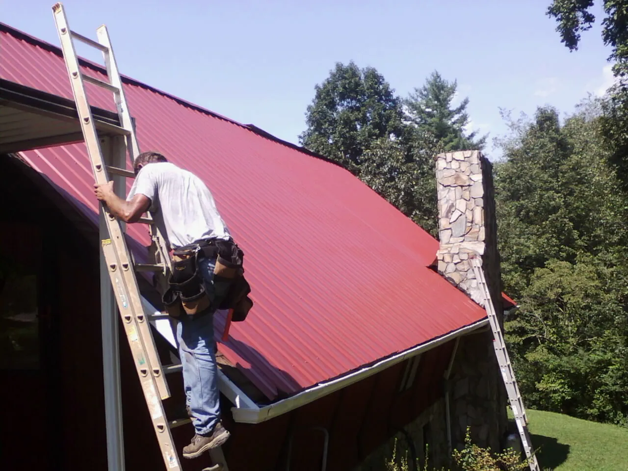 Roofer on a ladder beside a red metal roof and stone chimney — Soffit & Fascia Repair by Roofing Brothers in Millheim